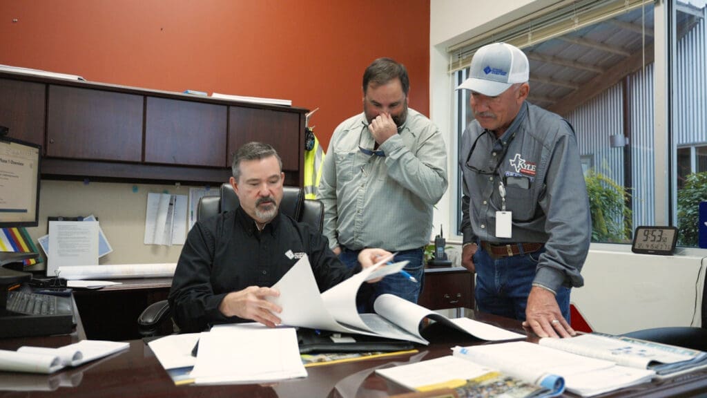 Three members of the Public Works Department including Street Superintendent Jesse Shroyer review documents together at a desk; one is seated and showing paperwork, while the other two stand attentively, looking at the papers. A large window lets in sunlight from outside.