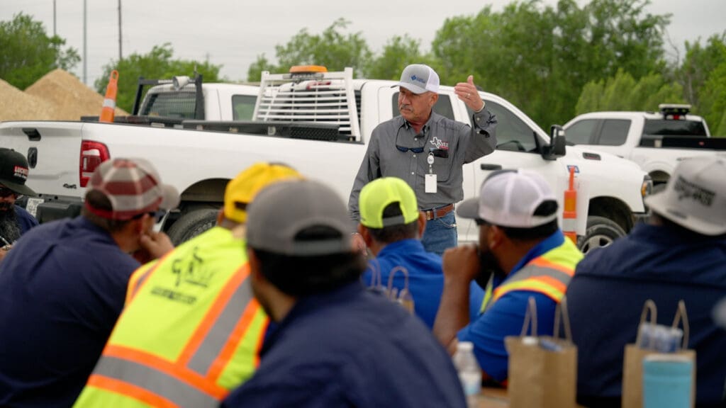 Streets Superintendent Jesse Shroyer speaks to a group of workers wearing safety vests and caps, seated outdoors near parked white trucks and construction materials.