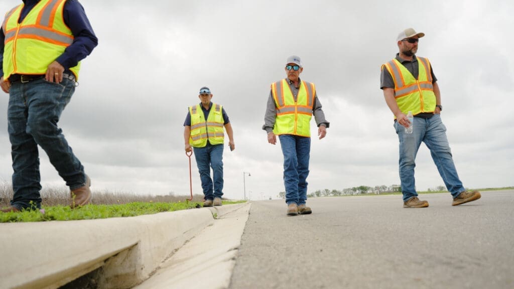 Four construction workers wearing yellow safety vests and jeans walk along a road under a cloudy sky. One worker carries a shovel, and another holds a bottle. They walk near a concrete curb and grassy area.