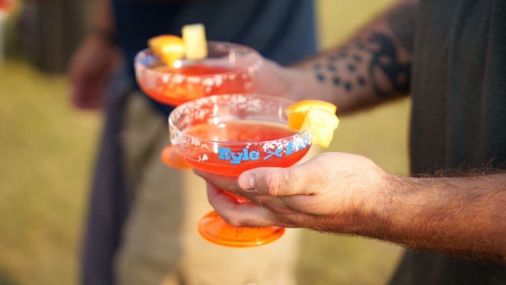 A person with tattooed arms holds two orange margarita glasses filled with a red drink, garnished with orange and pineapple slices, and a salted rim. Another person stands in the background.