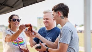 Three people smiling and holding barbecue ribs at an outdoor event, with two of them about to clink their ribs together in a toast. They are standing under a tent and appear to be enjoying themselves.