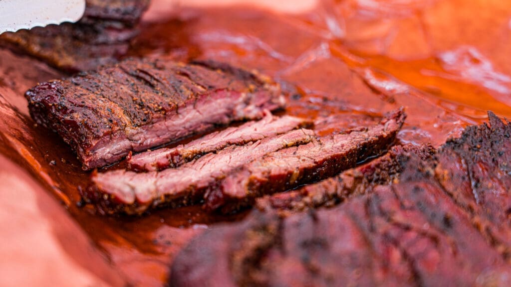 Close-up of sliced, smoked brisket on a sheet of reddish butcher paper, showing a dark, seasoned outer crust and juicy, tender meat inside.