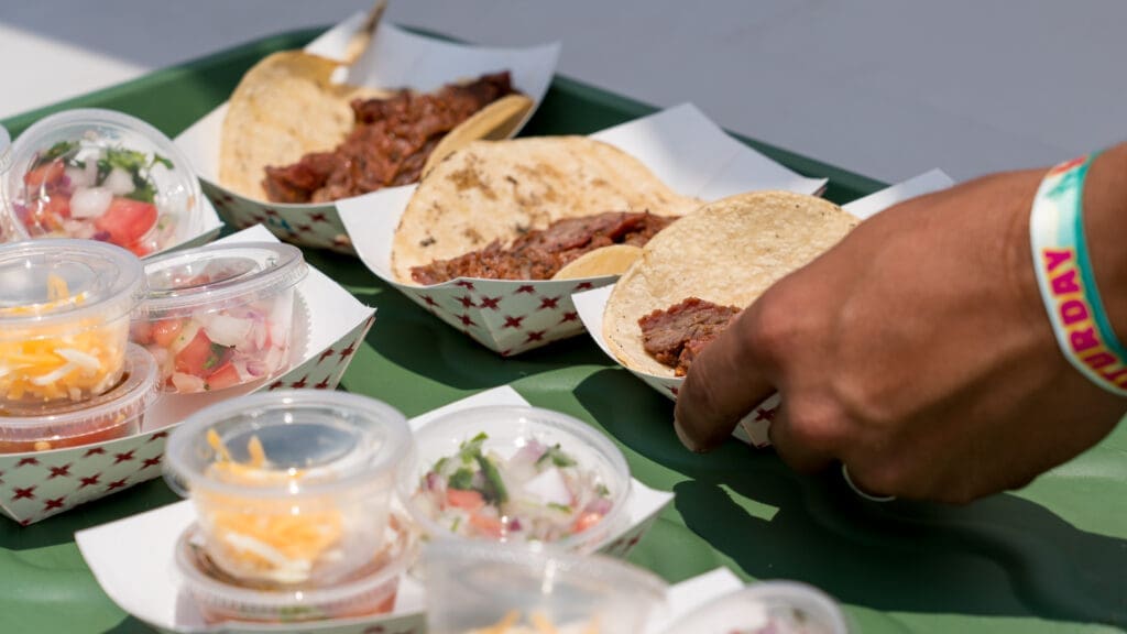A person’s hand picks up a taco from a tray holding several tacos in paper containers, accompanied by small plastic cups filled with toppings like salsa, pico de gallo, and shredded cheese.