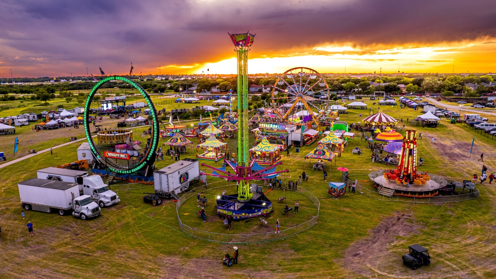A colorful carnival at sunset with rides, including a Ferris wheel, ring ride, and spinning tower, surrounded by people, tents, trucks, and grassy fields under a dramatic sky.