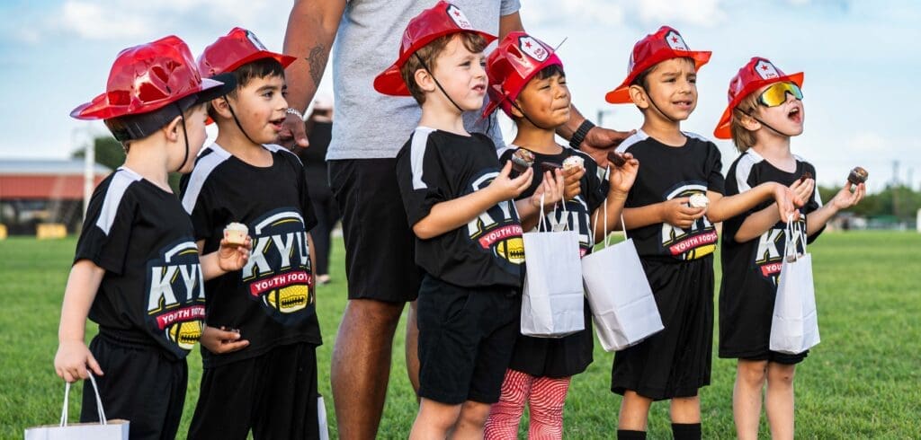 Six young children in black sports uniforms and red plastic firefighter hats stand in a row on grass, holding snacks and white bags, smiling and laughing, with an adult behind them. It's a bright, sunny day.