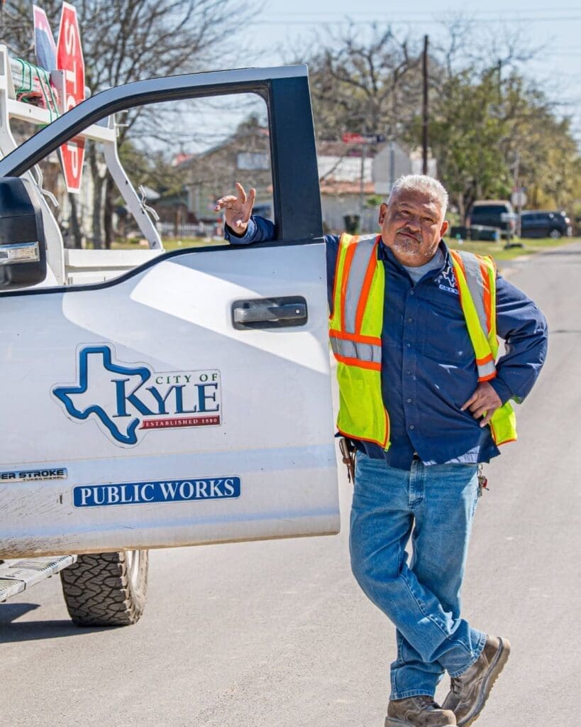 A City of Kyle Public Works employee in a safety vest leans against an open door of a white City of Kyle Public Works truck, standing on a sunny street with trees and houses in the background.