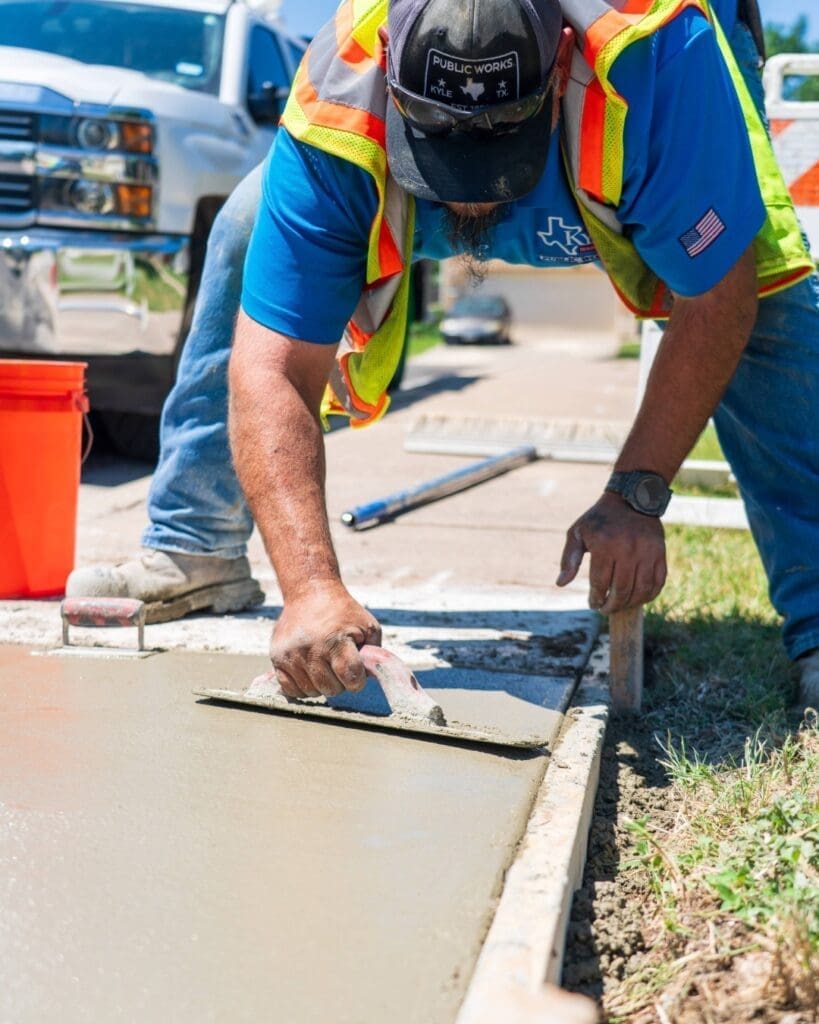A City of Kyle Publics Works employee in a safety vest smooths wet concrete on a sidewalk with a trowel, while kneeling next to a curb. A bright orange bucket and a truck are visible in the background.