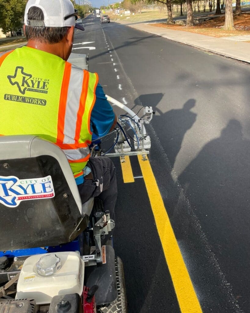 A City of Kyle Public Works employee wearing a "City of Kyle Public Works" vest operates a machine to paint fresh yellow lines on a newly paved road under clear skies.