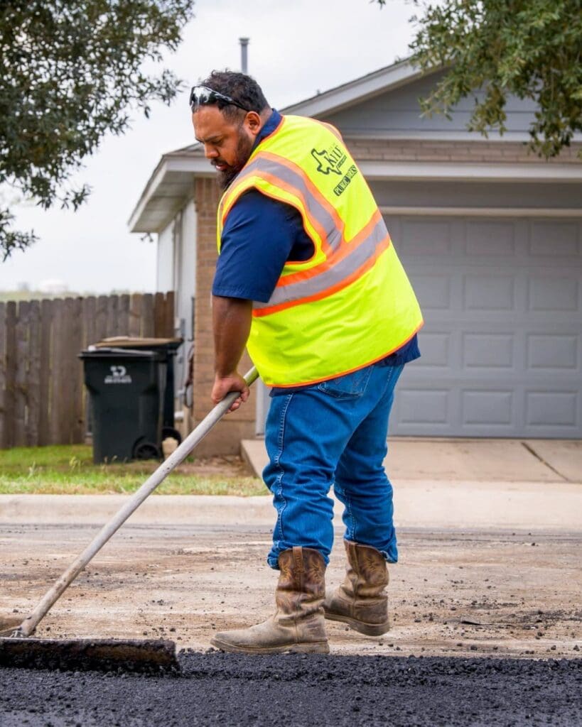 A City of Kyle Public Works employee in a yellow safety vest and boots spreads asphalt on a residential street with a rake. A house, driveway, and trash bin are visible in the background.