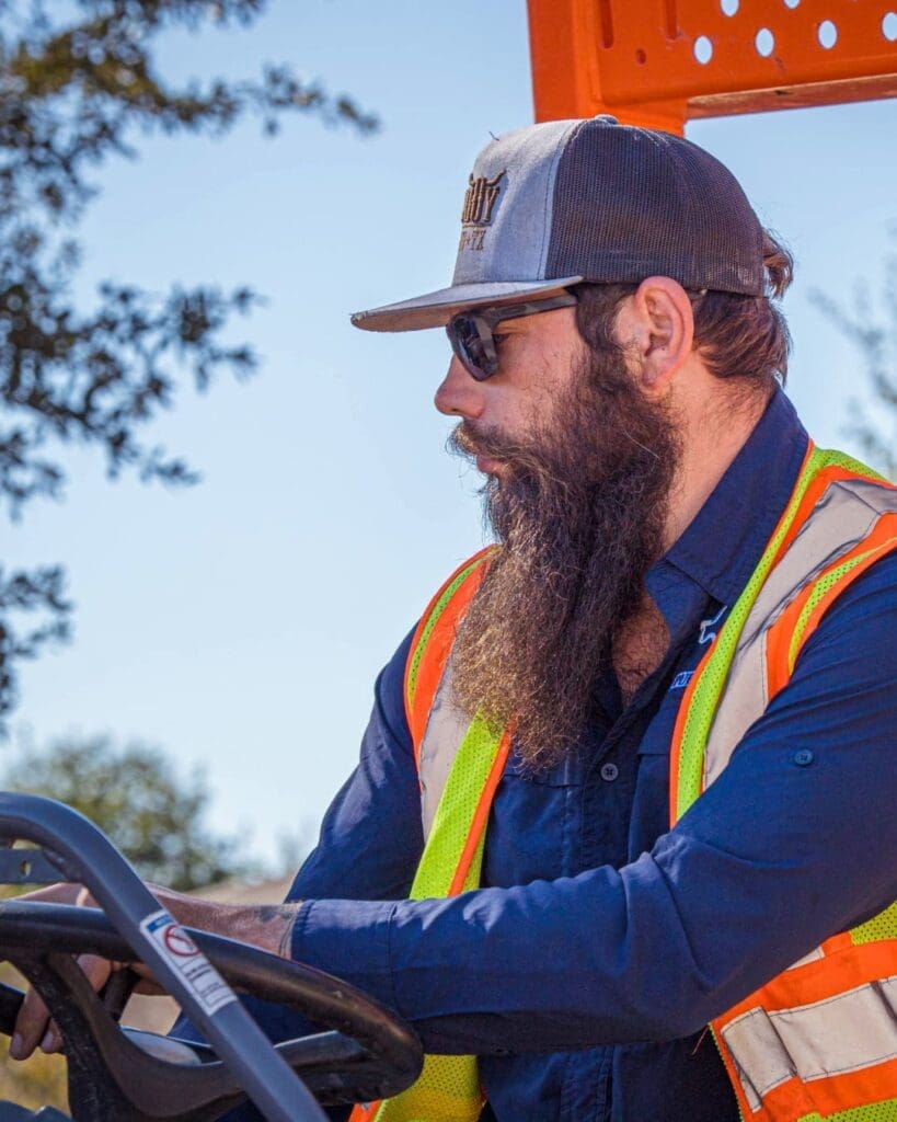 A City of Kyle Public Works employee wearing sunglasses, a trucker hat, and a reflective safety vest operates heavy machinery outdoors on a sunny day, with trees and blue sky in the background.