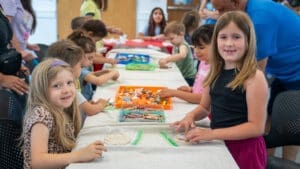 A group of young children sit and stand around a long table covered with art supplies, such as crayons and markers. Two girls in the front smile at the camera while others focus on their creative activities.