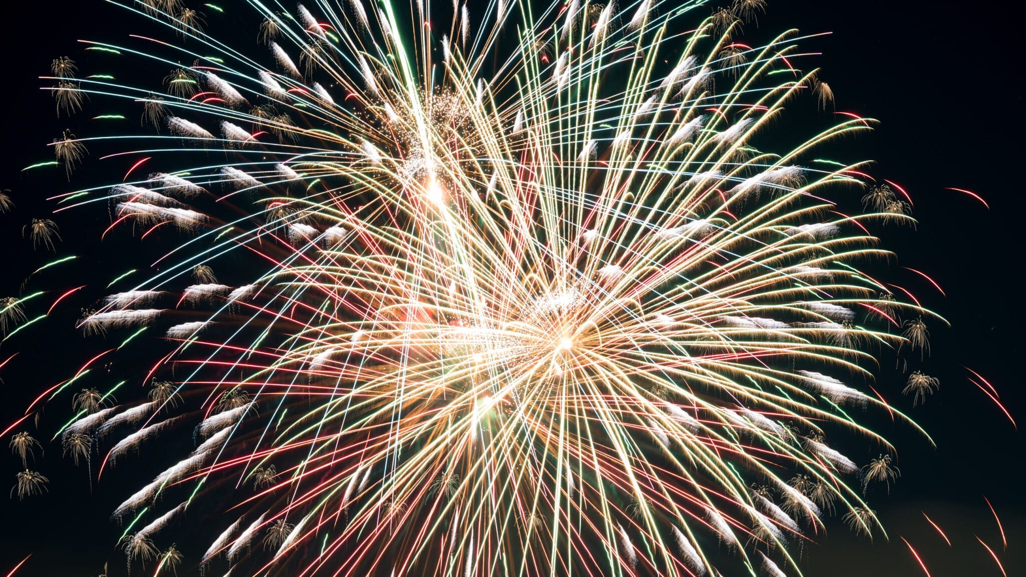 Colorful fireworks burst in the night sky, displaying streaks of red, white, and green light radiating outwards from a central point, creating a vibrant and celebratory scene.
