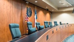 A modern courtroom with five empty black chairs behind a curved wooden bench, American and state flags, and a large official seal on the wood-paneled wall.