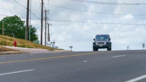 A black Jeep drives uphill on a wide, empty road with power lines overhead. There's a fire hydrant, grassy slope, and trees on the left, under a partly cloudy sky.