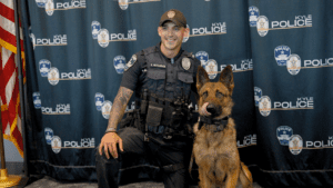 A smiling police officer kneels next to Atlas, a Malinois/German Shepherd police dog in front of a backdrop with "Kyle Police" logos.