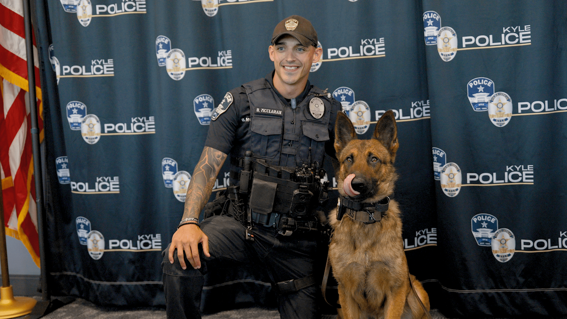 A smiling police officer kneels next to Atlas, a Malinois/German Shepherd police dog in front of a backdrop with "Kyle Police" logos.