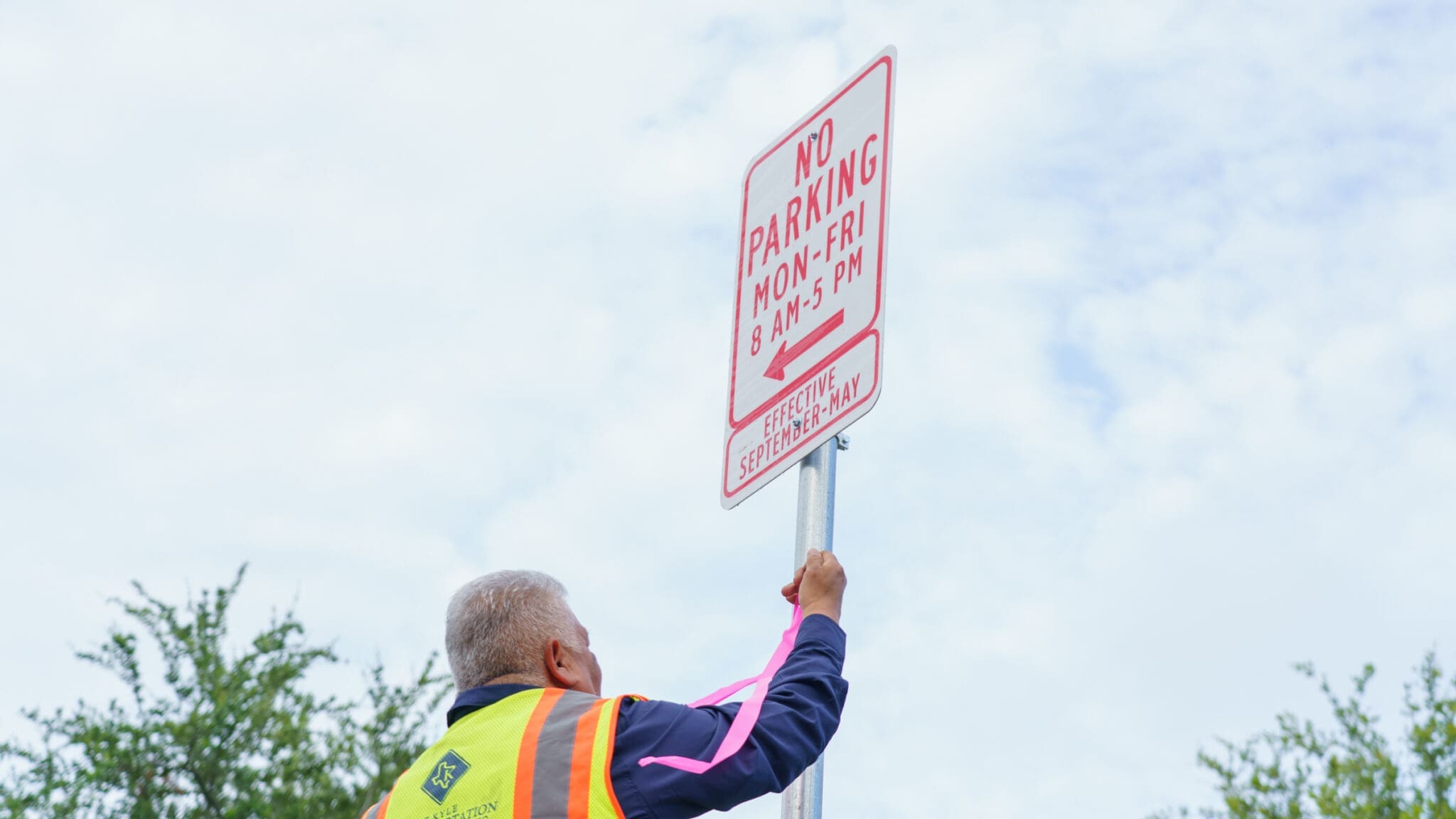 A worker in a safety vest installs a "No Parking Mon-Fri 8 AM-5 PM" sign on a pole outdoors, with trees and a cloudy sky in the background.