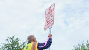 A worker in a safety vest installs a "No Parking Mon-Fri 8 AM-5 PM" sign on a pole outdoors, with trees and a cloudy sky in the background.