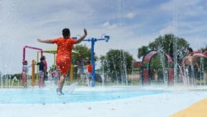 A child in an orange outfit runs joyfully through water jets at an outdoor splash pad, with other children playing in the background under a sunny sky.