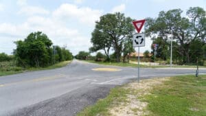 A rural roundabout with a yellow center is shown at the intersection of Roland Lane and South Main Street, surrounded by trees and grass. A yield sign and roundabout traffic sign are posted at the entrance. The sky is partly cloudy.