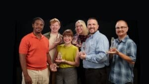 Six smiling people stand together holding clear glass awards, dressed in casual and business-casual clothing, against a plain black background.