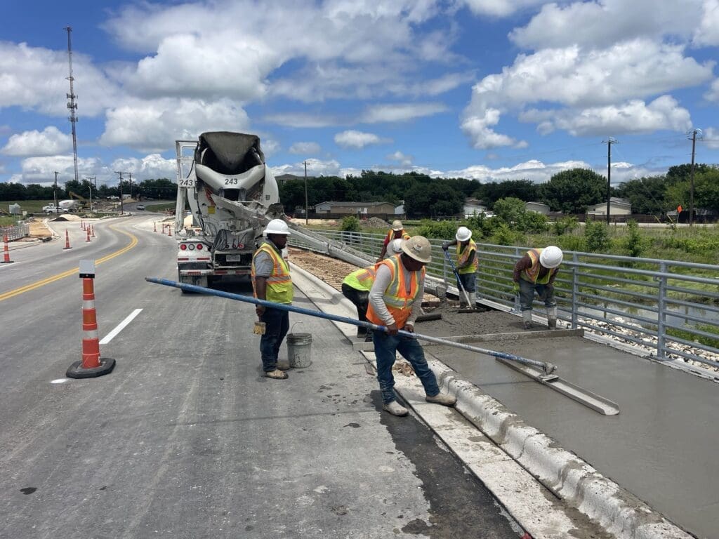 Several construction workers wearing orange safety vests and hard hats smooth out concrete along the Kohlers to Seton extension in Kyle, Texas. 