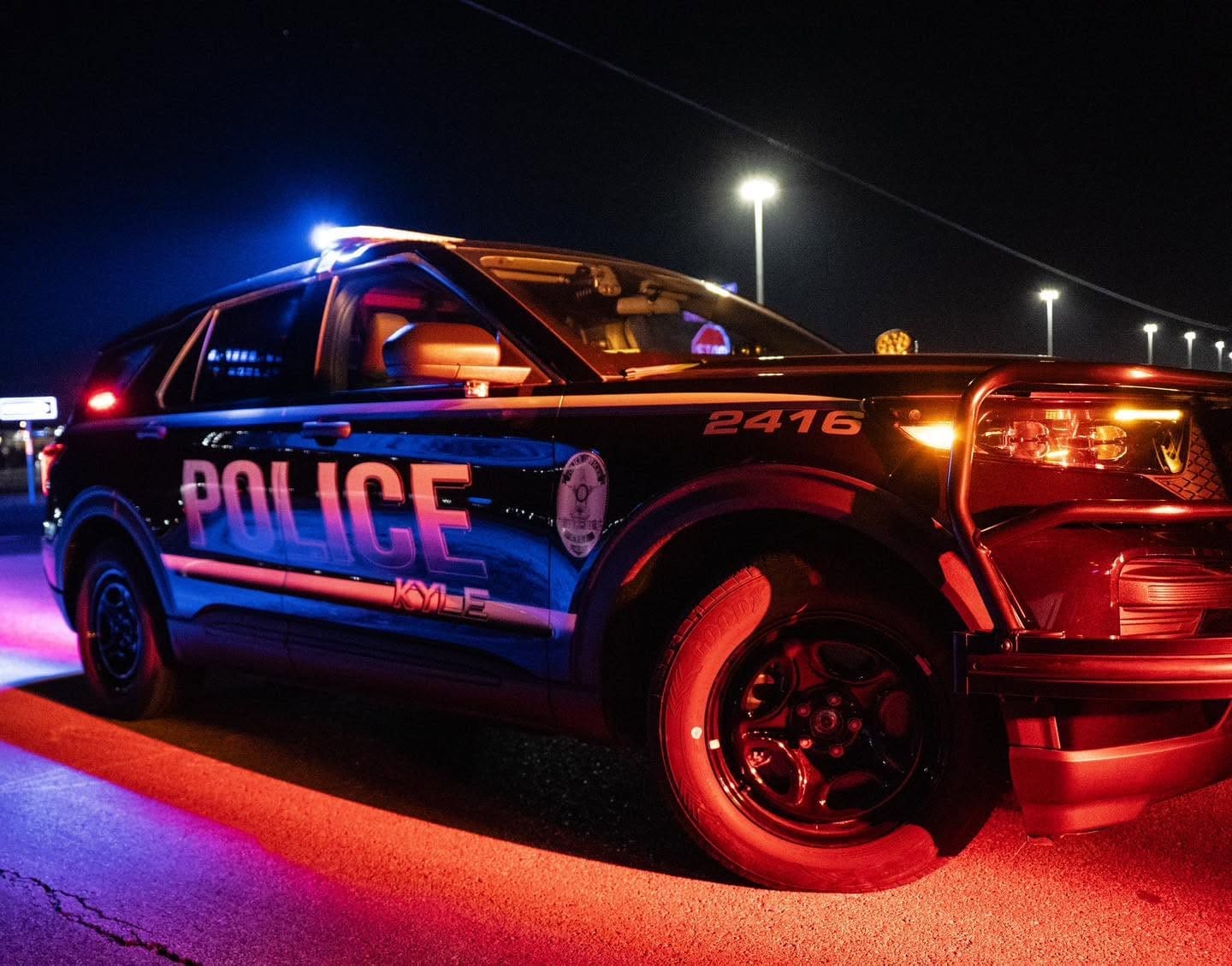 A police SUV with flashing red and blue lights is parked on a road at night. Streetlights are visible in the background, and the vehicle's side says "POLICE" in large letters.