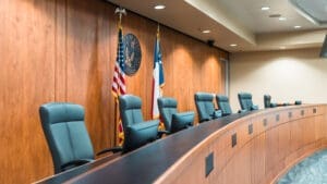 A row of empty leather chairs sits behind a long wooden desk in a courtroom, with the U.S. and Texas flags and an official seal displayed on the wood-paneled wall.