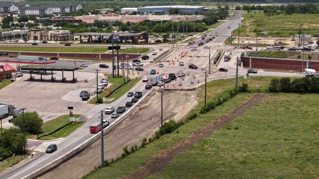 A drone photograph of several cars waiting at a stoplight at the intersection of Windy Hill Road and Interstate 35 in Kyle, TX. To the right of the intersection a large tract of land is cleared and several traffic cones can be seen. 