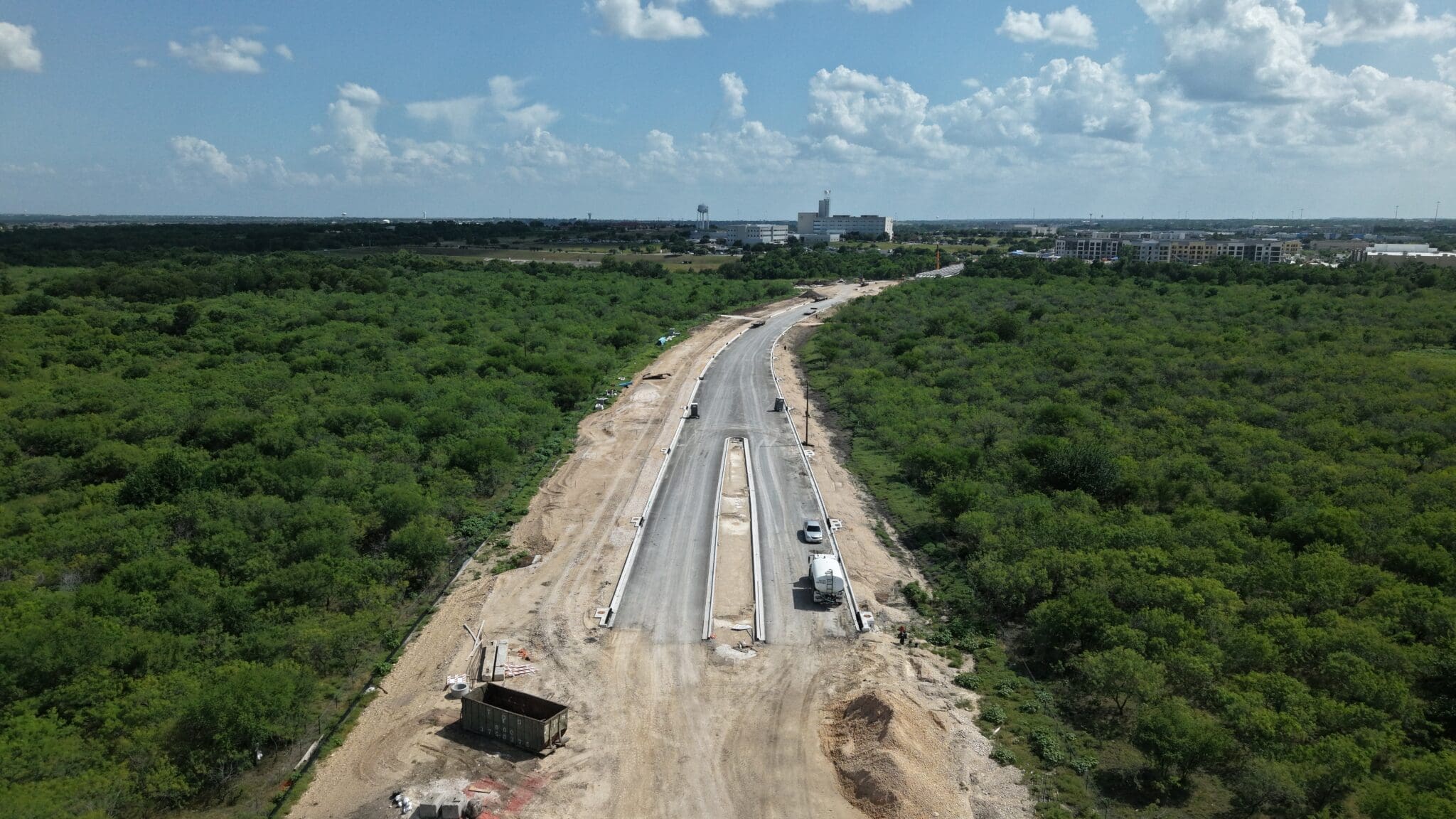Aerial view of the partially constructed Kohlers to Seton extension cutting through dense green vegetation, with construction vehicles and equipment visible. Buildings and a water tower appear in the distant background under a partly cloudy sky in Kyle, Texas.