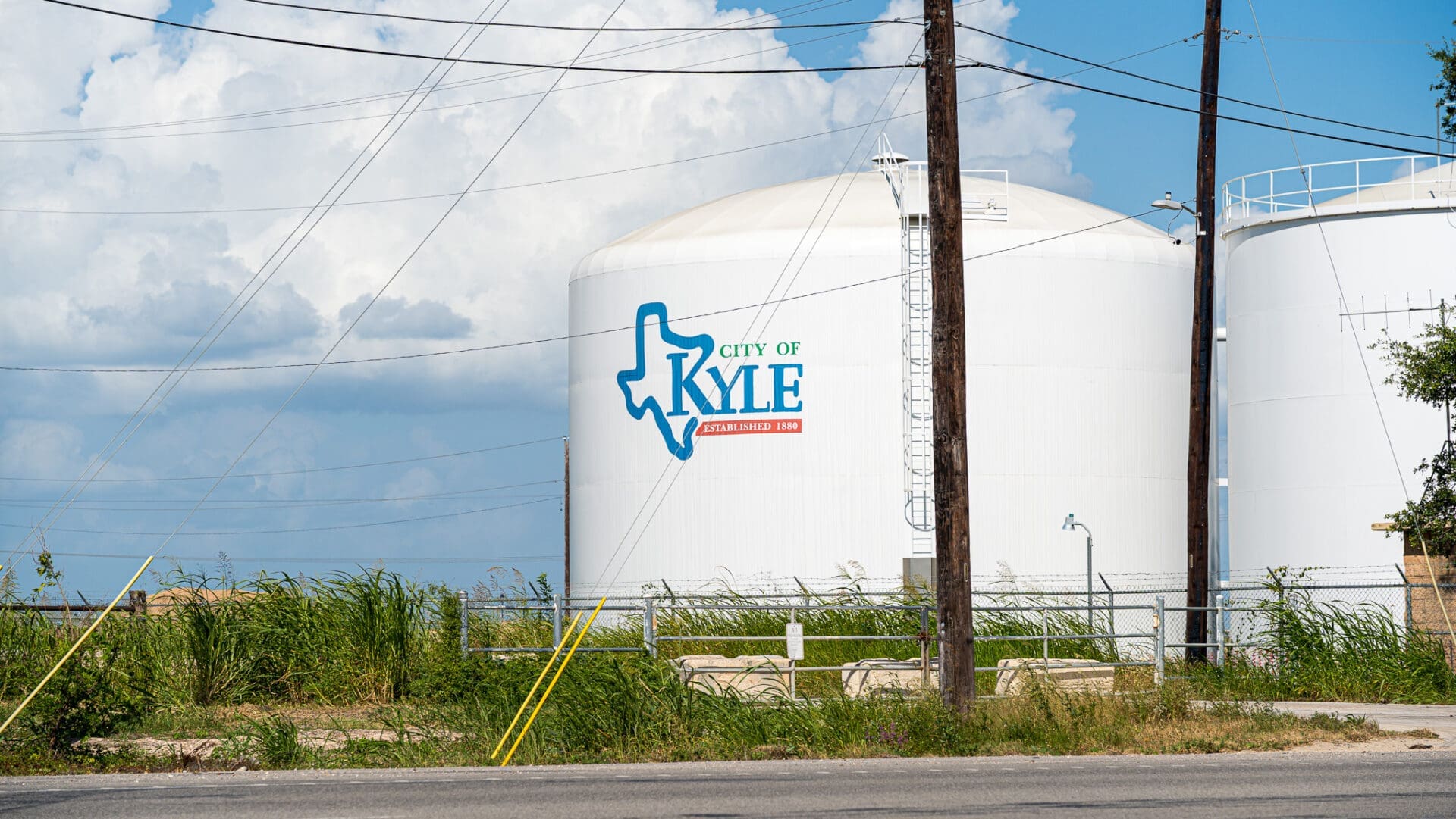 White water tank with "City of Kyle" and a Texas outline logo, next to another tank, behind grass, power poles, and a fence under a blue sky with clouds.