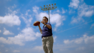 Brooke Collins, the City of Kyle's Recreation Coordinator, holds a football, preparing to throw it, with a bright blue sky, scattered clouds, and stadium lights in the background in Kyle, TX.
