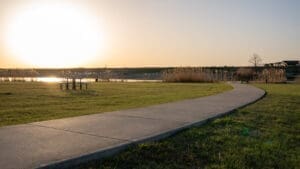 A winding concrete path leads past a picnic table toward a lake at sunset, with tall grasses and a clear sky in the background.